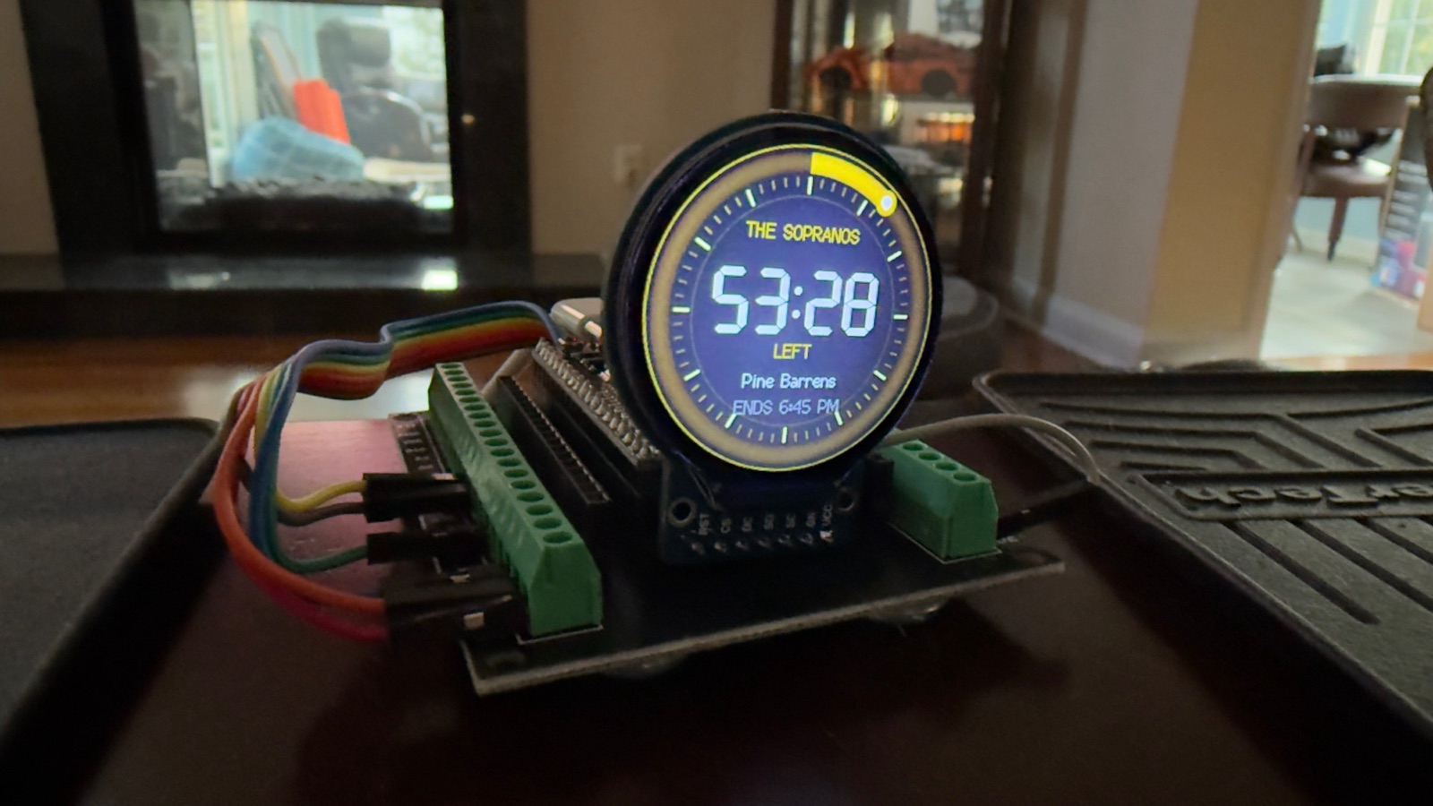 A round display on an ESP32 breakout board showing The Sopranos, 53:28 left, Pine Barrens, ends 6:45 PM